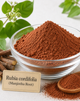 A clear glass bowl filled with reddish-brown powder labeled 'Rubia cordifolia (Manjistha Root)', with loose roots, a wooden spoon of the powder, and green leaves on a light background.