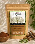 A stand-up pouch of Radico Organic Manjistha Powder stands on a light fabric surface, with a small wooden bowl of brown powder, loose root pieces, and green leaves in the background. The pouch label is visible and reads 'Radico Organic' and 'Manjistha Powder'.