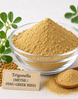 A clear glass bowl filled with tan powder, surrounded by green methi leaves, loose seeds, a wooden spoon with powder, and a small label reading 'Trigonella (METHI / FENU-GREEK SEED)'.