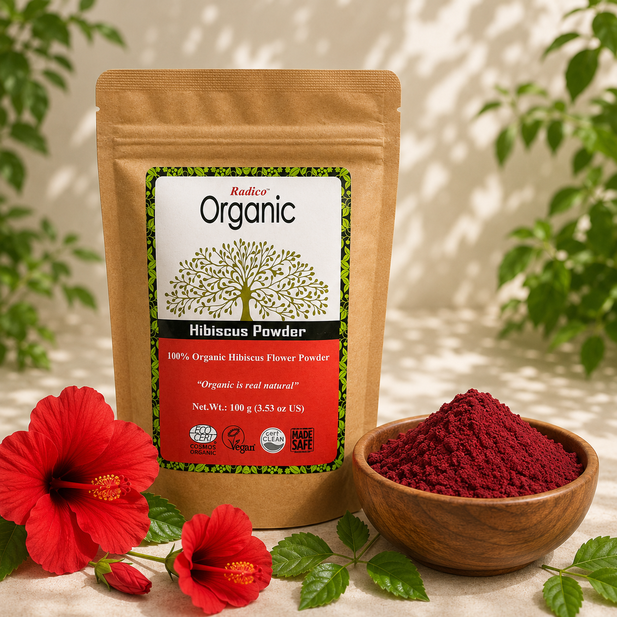 A stand-up pouch of Radix Organic Hibiscus Powder stands on a light surface with red hibiscus flowers and a small wooden bowl of the powder in front. The pouch label reads Organic and Hibiscus Powder, with green leaves and a softly blurred outdoor background.