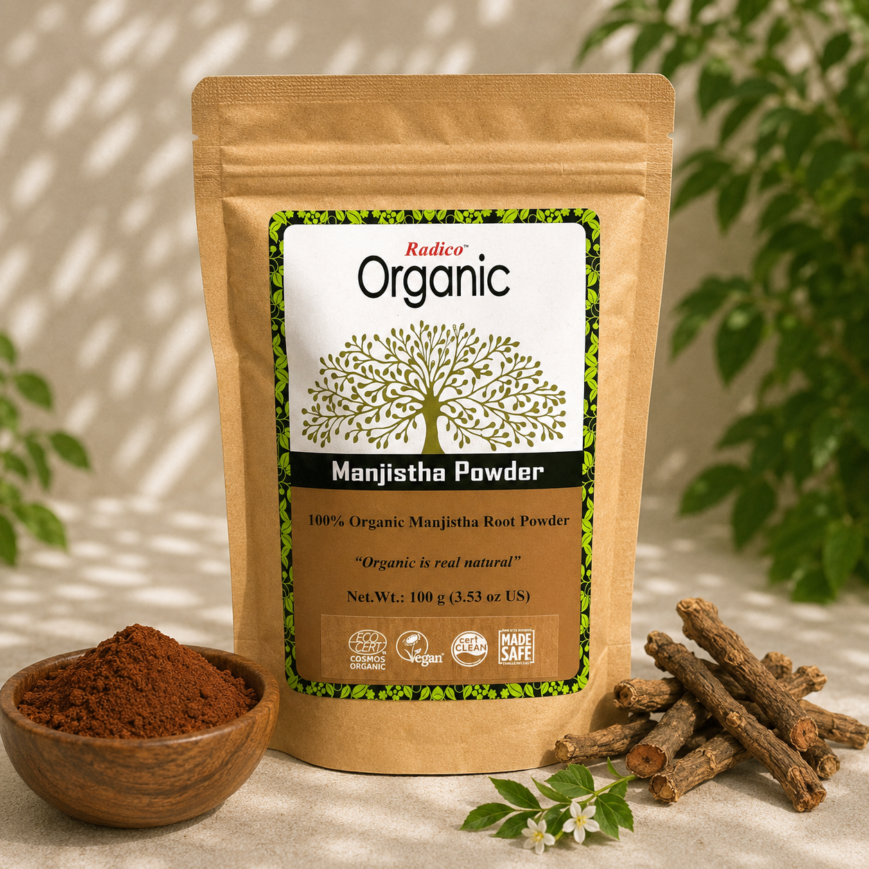 A stand-up pouch of Radico Organic Manjistha Powder stands on a light fabric surface, with a small wooden bowl of brown powder, loose root pieces, and green leaves in the background. The pouch label is visible and reads 'Radico Organic' and 'Manjistha Powder'.