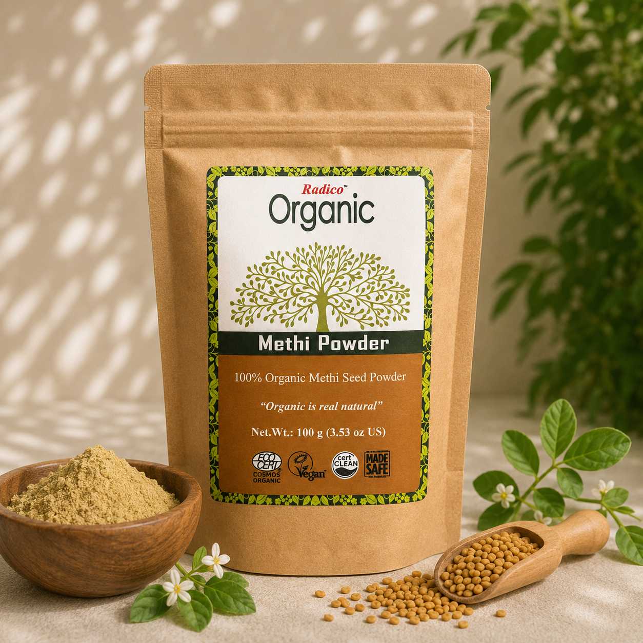 A stand-up kraft pouch of Radico Organic Methi Powder is shown on a light neutral surface with a bowl of loose powder, a wooden scoop of seeds, and green plant leaves in the background.